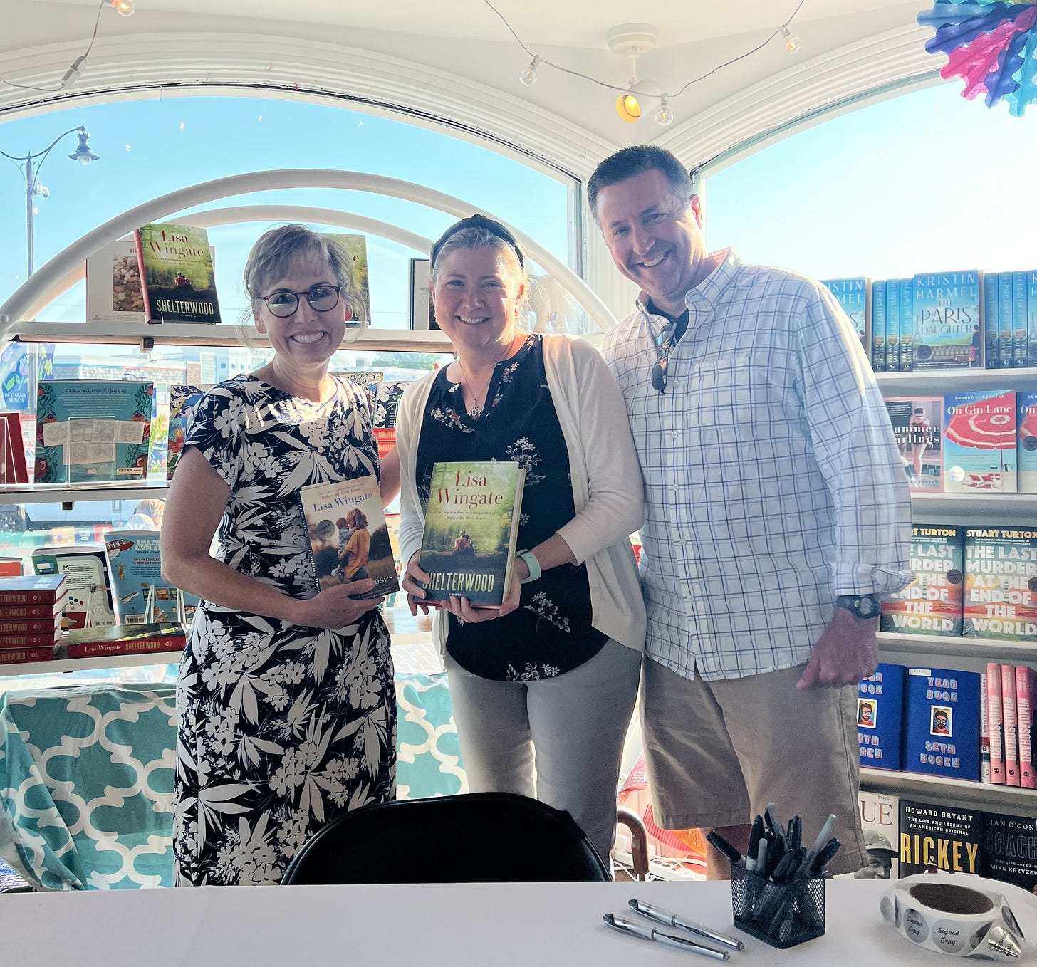 Lisa, Patty, and Adam standing behind a table for autographing, with bookshelves and arched windows in the background. Lisa and Patty are holding copies of Lisa’s books, Tending Roses and Shelterwood.
