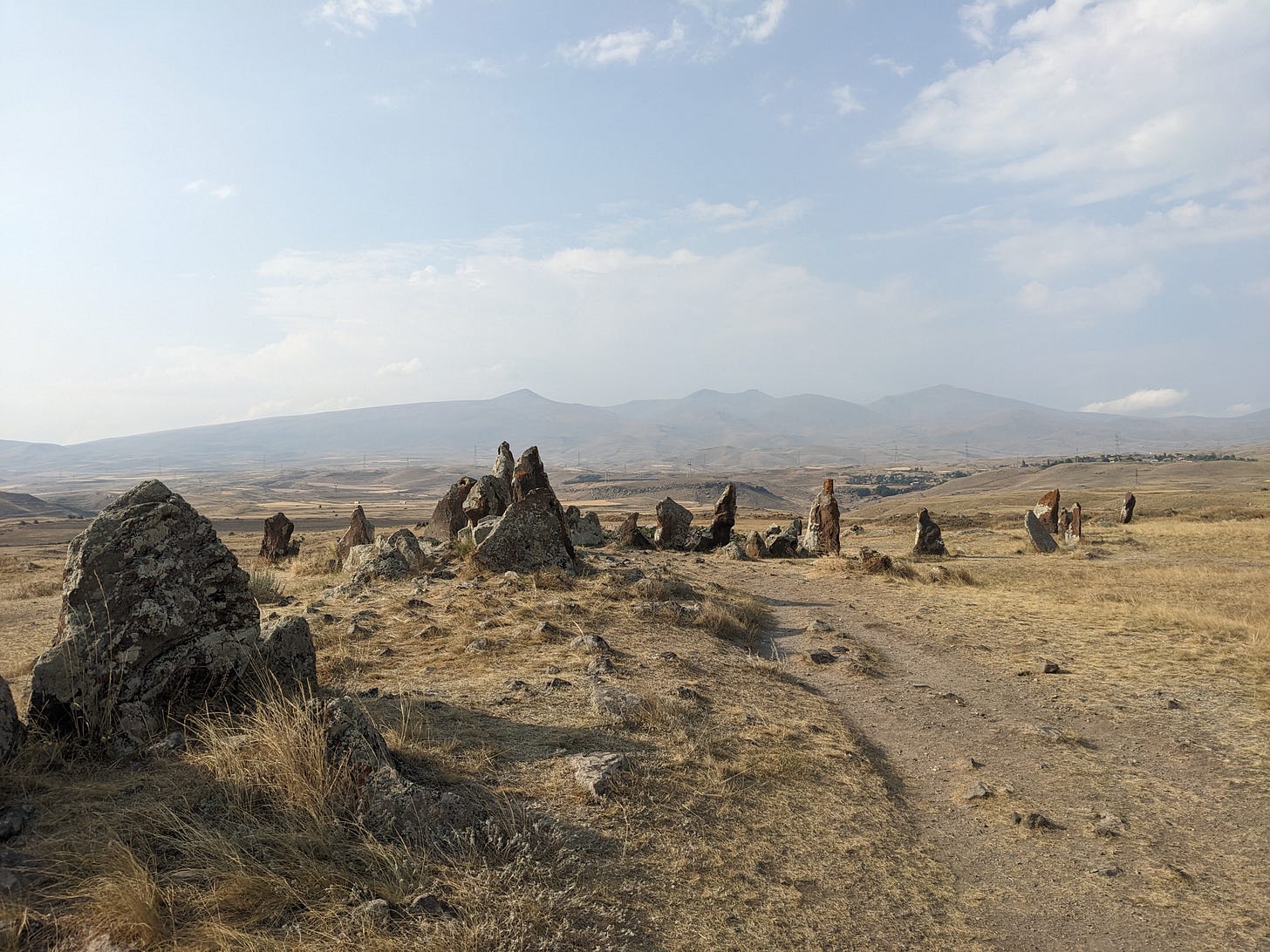 A series of jagged, grey stone stand in a line on a golden hillside. A blue sky is beyond and a dirt path is in the foreground.   