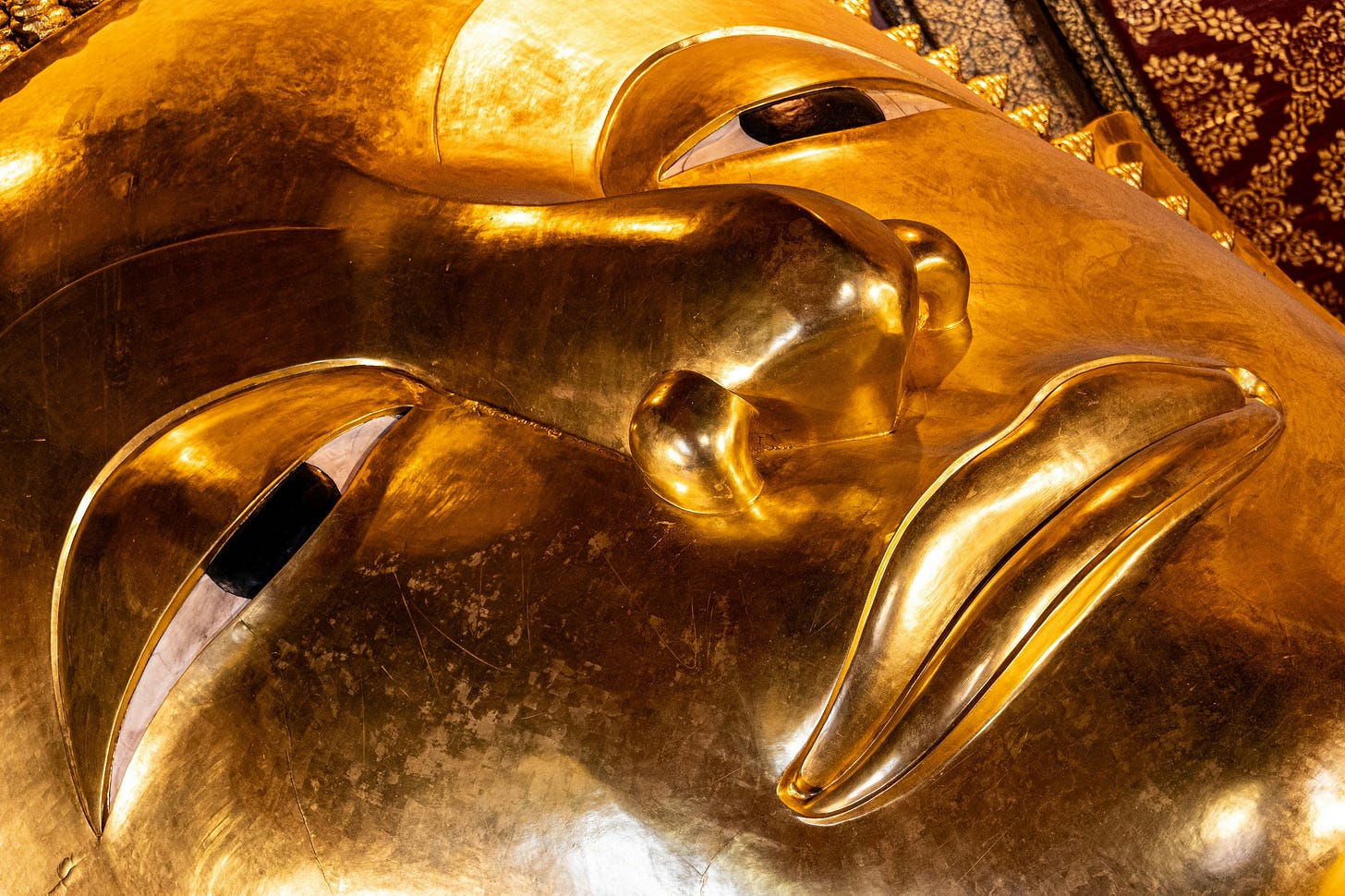 Close-up photograph of the face of a reclining golden Buddha statue, shot at an angle that makes the features almost abstract. The heavy-lidded eyes are half-closed. The surface is worn in places, ancient gold catching warm light.