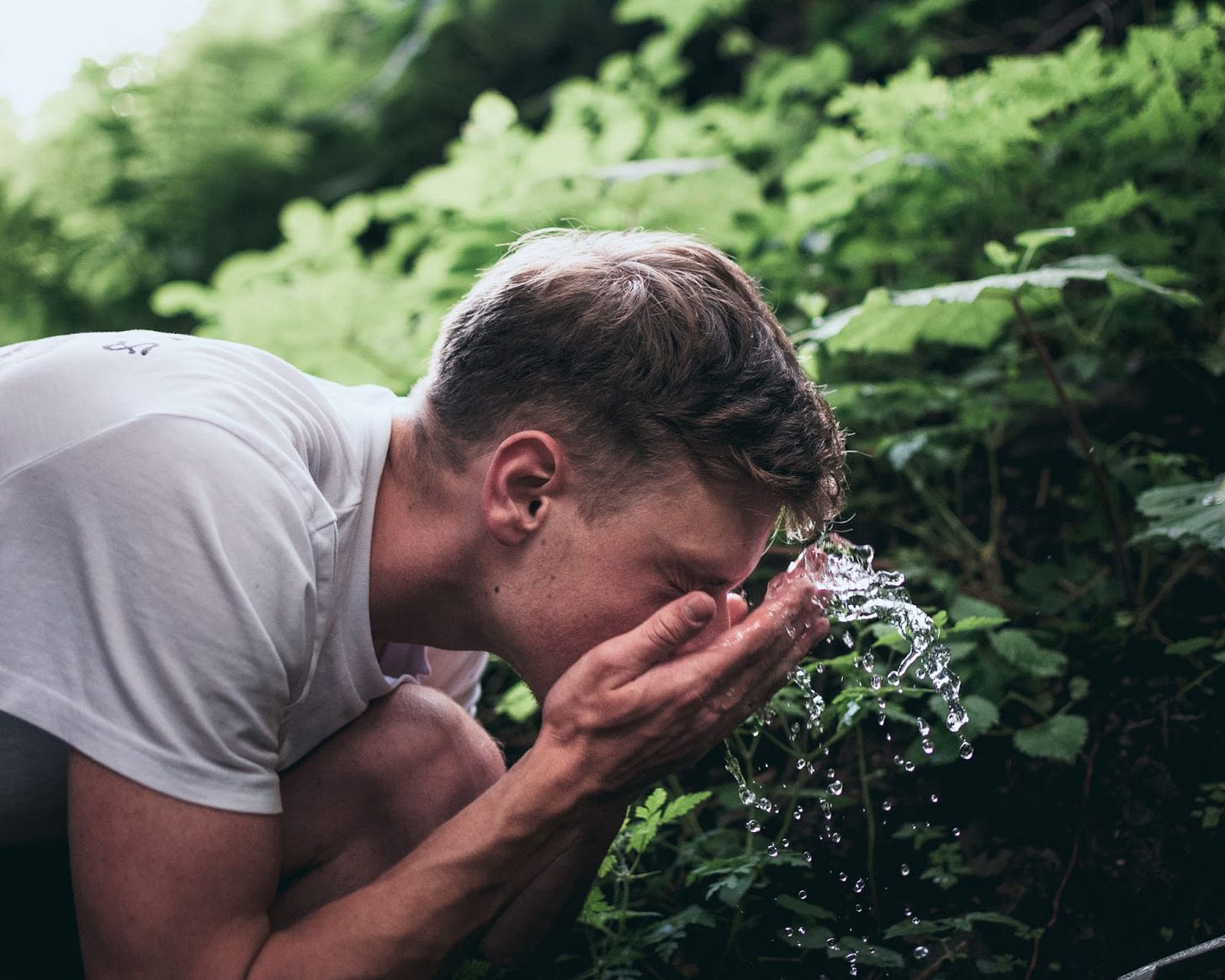 A man in a white t-shirt splashing his face with water in front of an out of focus green plant background. A man in a white t-shirt splashing his face with water in front of an out of focus green plant background.