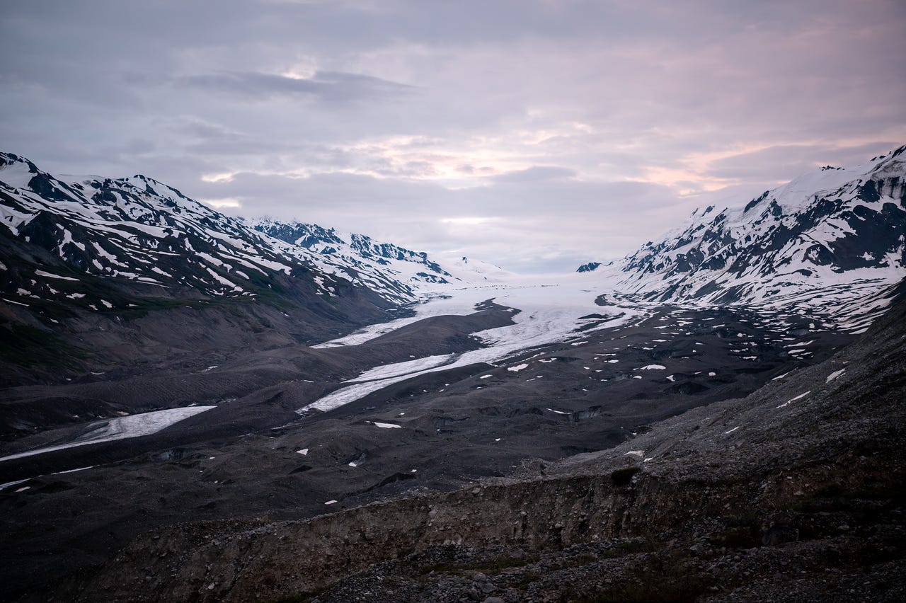 A broad glacier fills a mountain valley, its lower reaches buried under dark rock debris so that only stripes of white ice show through, widening to clean snow-covered ice upvalley. Snow-laced peaks rise on both sides, and the dawn sky glows pale pink and lavender through breaks in soft grey cloud. The light is the quiet, shadowless pink of an Alaska summer sunrise.