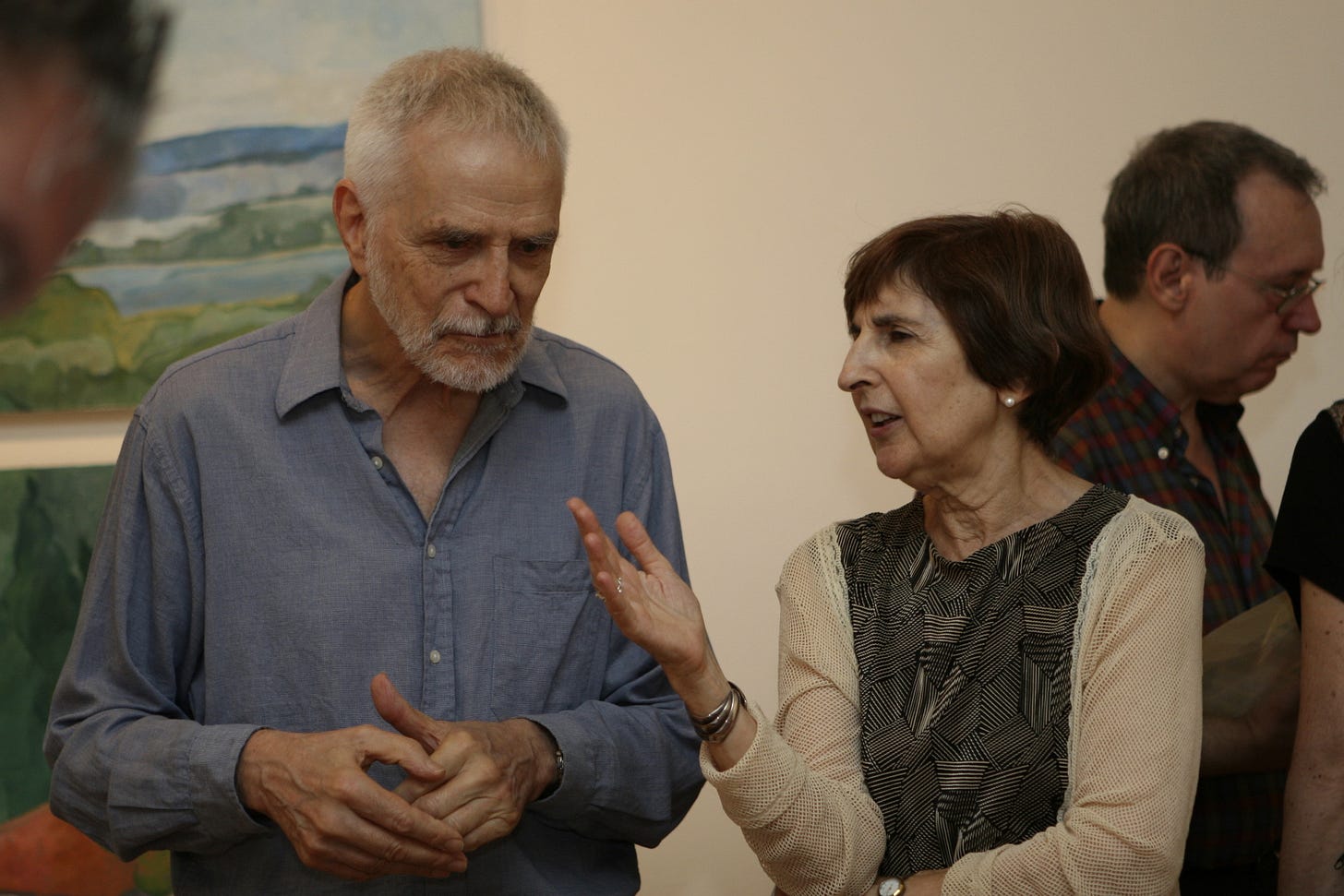 An older man and woman in conversation at an art gallery with other people surrounding them, and paintings on the wall behind.