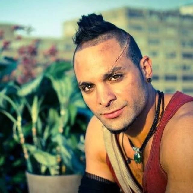 Close-up portrait of Vaas Montenegro, played by Michael Mando; a muscular man with a shaved-sides mohawk and a thin scar across his forehead, wearing a red tank top and layered necklaces. He looks directly at the camera with an intense, unsettling expression, while tropical plants and blurred buildings appear in the background under bright daylight.