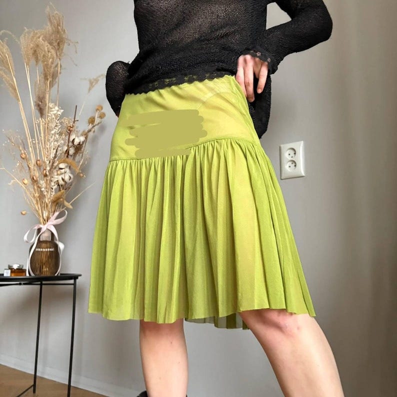 May include: A lime green pleated skirt with a gathered waist. The skirt is knee-length and appears to be made of a lightweight fabric. The model is wearing a black long-sleeved top.
