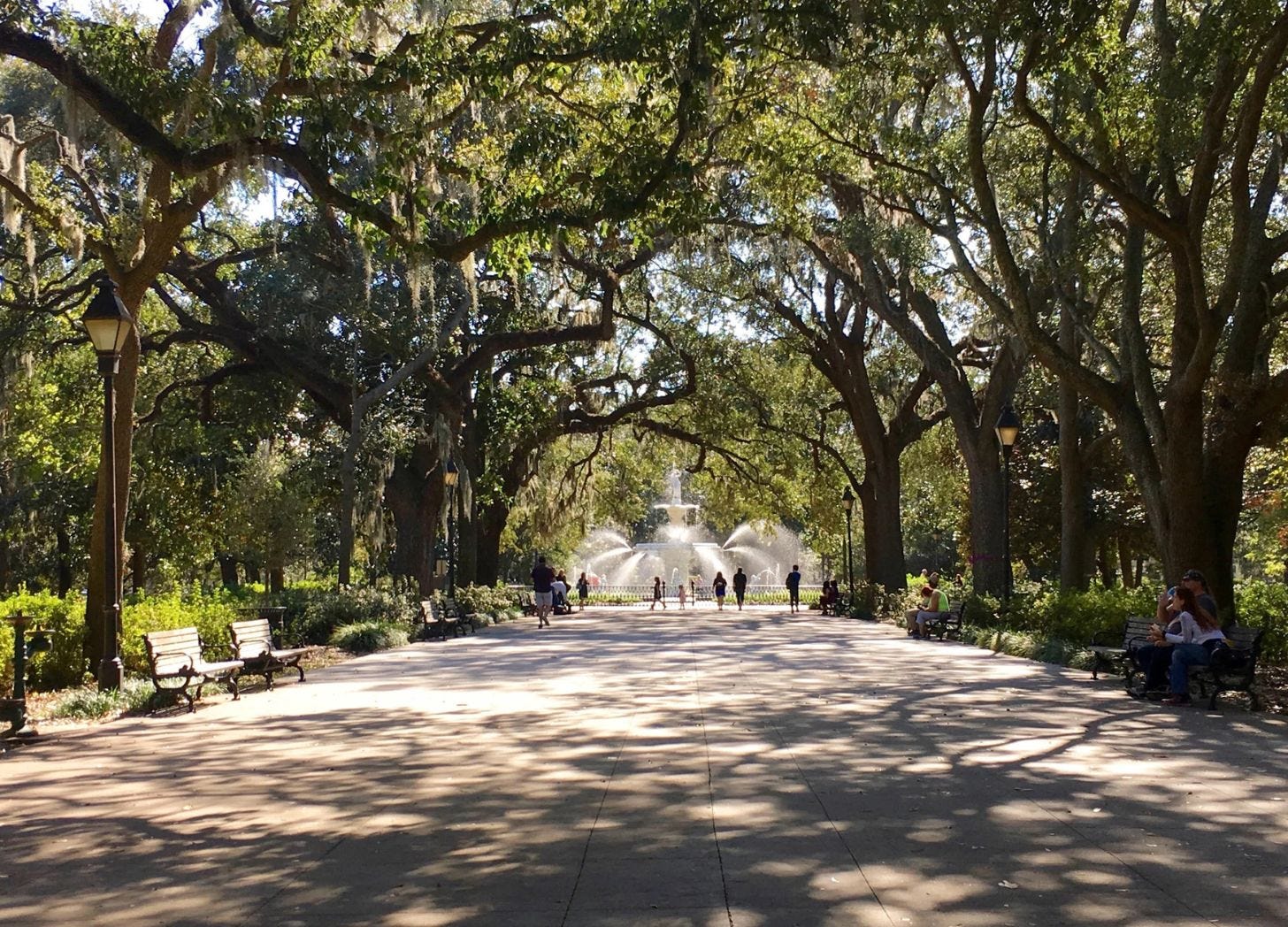 People walking through a shaded Savannah square lined with live oak trees, benches, and a central fountain. People walking through a shaded Savannah square lined with live oak trees, benches, and a central fountain.