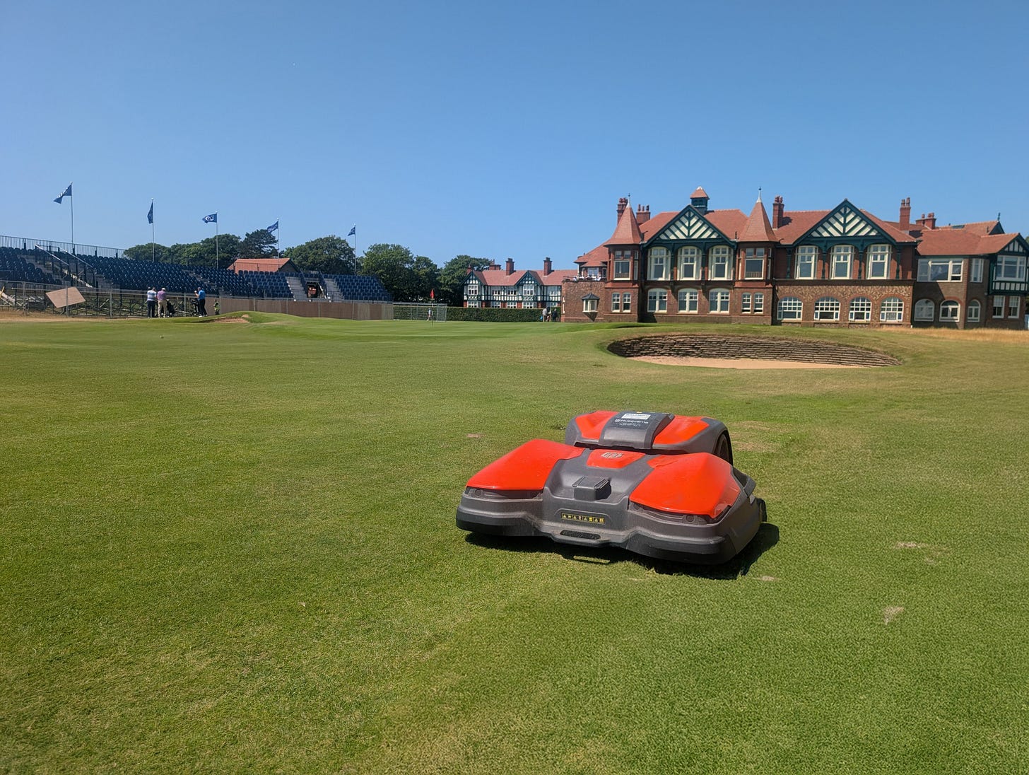 Two Husqvarna robotic mowers on the closing green area at Royal Lytham & St Annes, with championship grandstands and the Tudor clubhouse behind.