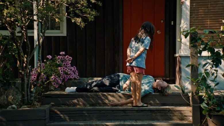 In a still from the film "Blue Heron," a young girl stands over her brother, who is laying on the ground, outside of their front door. 