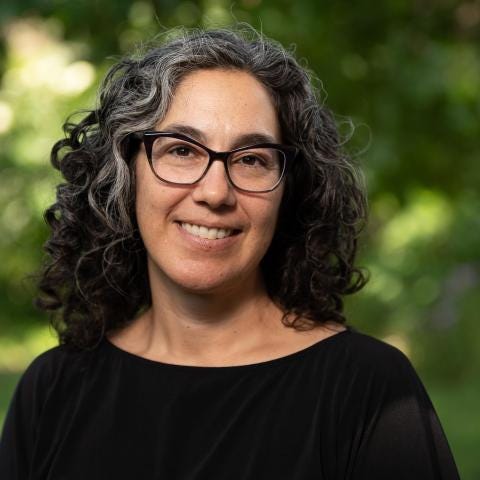 A head-and-shoulders portrait shows a person with shoulder-length gray curly hair wearing dark-framed glasses and a black top. The person is smiling slightly and standing outdoors in front of a softly blurred green background of trees or foliage, creating a natural, evenly lit setting.
