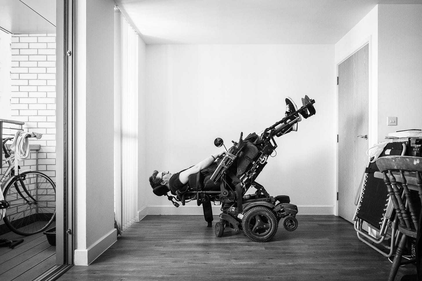 Jamie Hale in a bare living room, a deckchair on one side, a bike on the other. They are in an electric wheelchair, but tilted upside down, head near the floor, legs up in the air