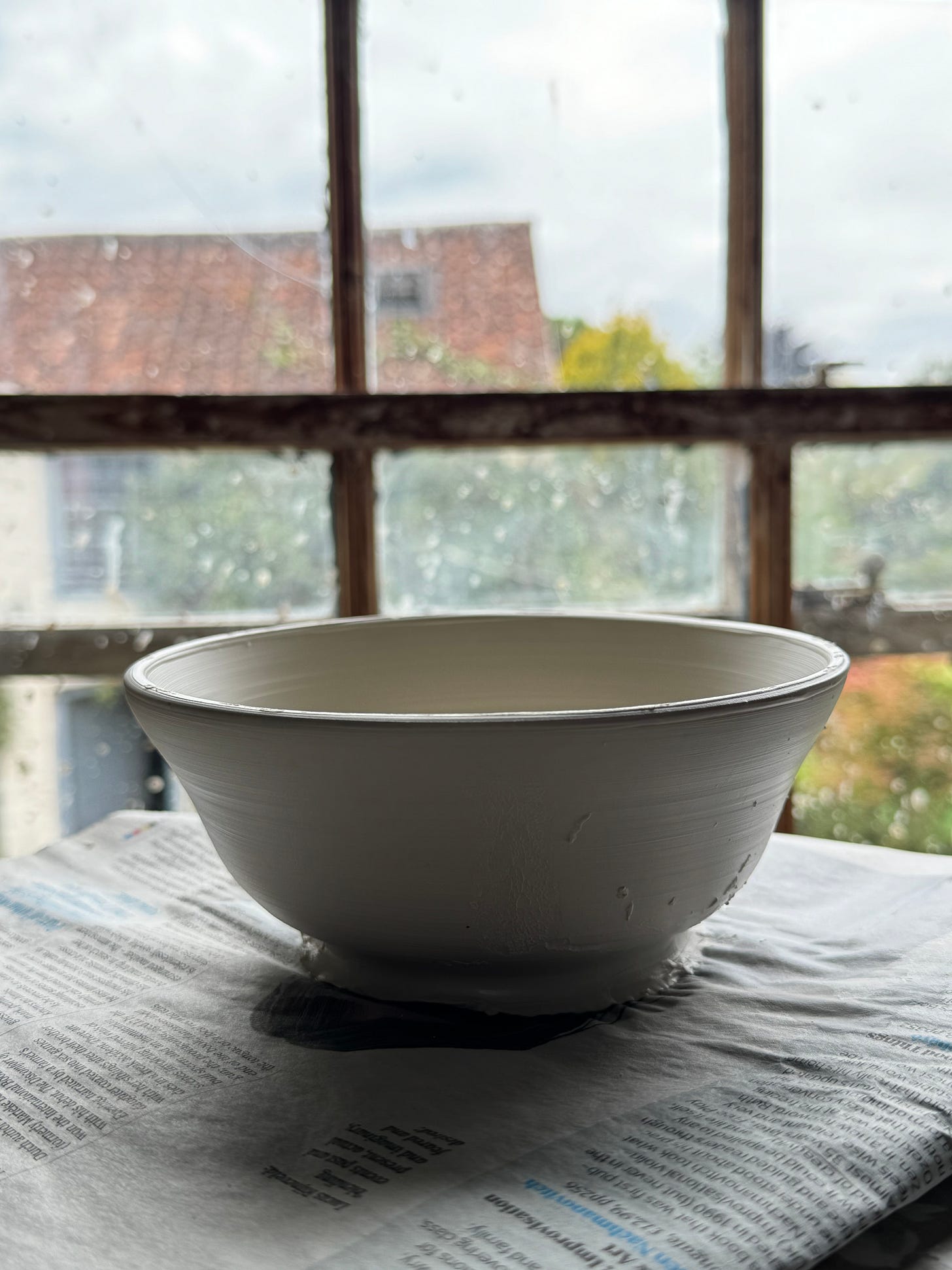 A freshly thrown porcelain bowl on a piece of newspaper in front of a wooden sash window