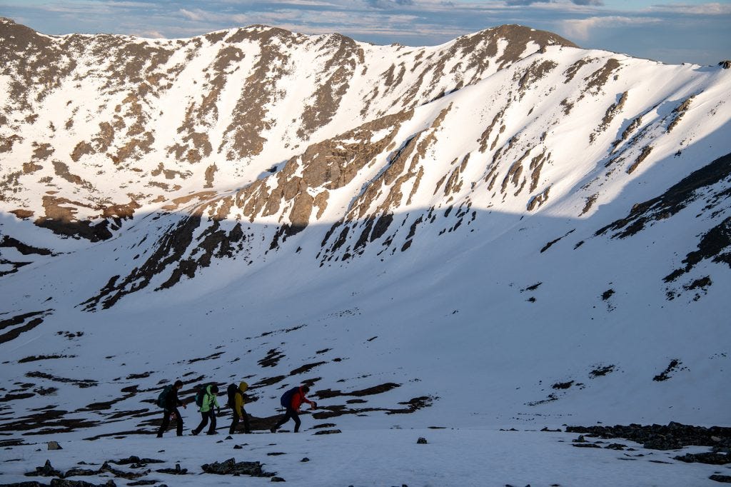 Four small figures cross a vast snowfield below a large snow-covered cirque headwall in evening light, dwarfed by the slope above them.