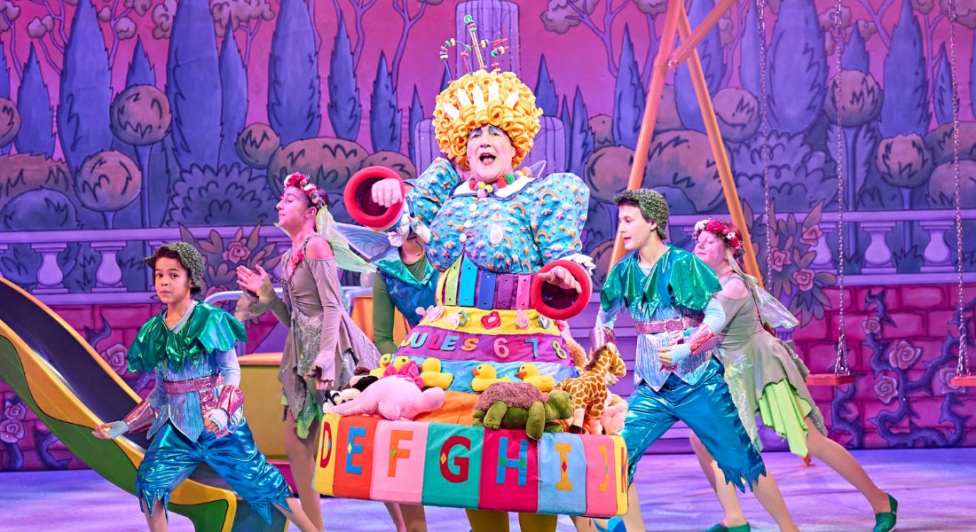 A man is standing centre stage wearing an elaborate, colourful dress which is designed with different children's toys on it. His mouth is open as he is mid singing. Behind him are four of the youth chorus dressed as fairies.