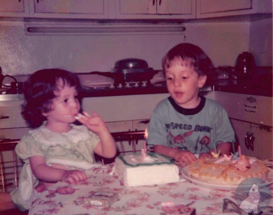 Vintage color photograph of two young children at a kitchen table with a floral tablecloth. On the left, a toddler-aged girl with curly dark hair in a pale green lace-collared dress licks frosting from her finger. On the right, a slightly older boy in a Speedy Bunny T-shirt sits in front of two birthday cakes: a smaller white sheet cake with a single lit candle and a larger round cake with several unlit candles. A 1970s-era kitchen with white cabinets is visible behind them. Vintage color photograph of two young children at a kitchen table with a floral tablecloth. On the left, a toddler-aged girl with curly dark hair in a pale green lace-collared dress licks frosting from her finger. On the right, a slightly older boy in a Speedy Bunny T-shirt sits in front of two birthday cakes: a smaller white sheet cake with a single lit candle and a larger round cake with several unlit candles. A 1970s-era kitchen with white cabinets is visible behind them.