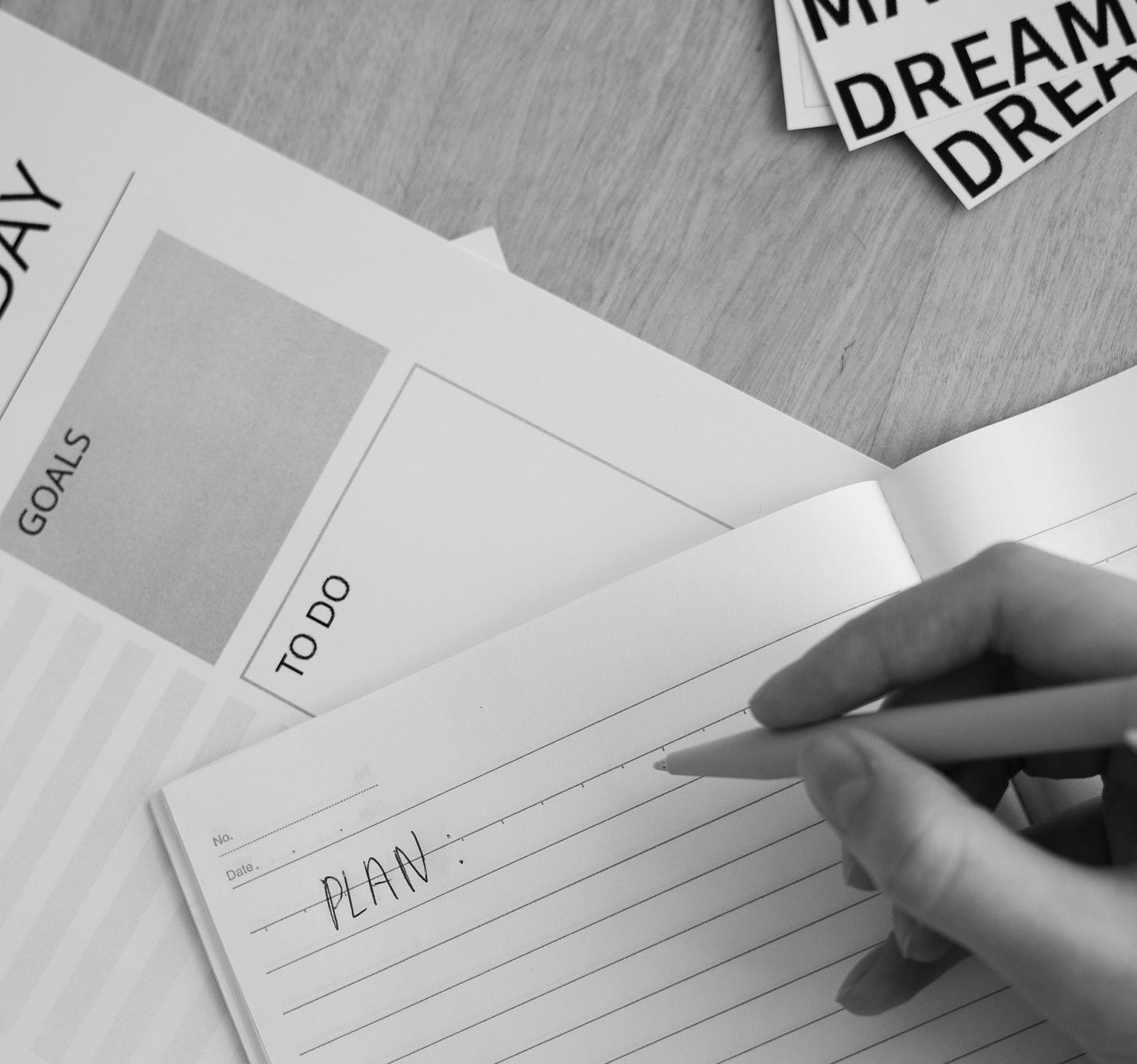 Black and white photo, overhead shot of a desk. A hand grips a pen mid-stroke, just finishing the word "PLAN" at the top of a lined notebook page. Underneath and fanned out is a daily planner showing GOALS and TO DO sections plus a time-blocked schedule running from 12:00 through 18:00, with dates 12 to 21 printed along the bottom edge. Top right corner has torn paper cutouts spelling out MAKE DREAMS, stacked at an angle like they were just pulled off something. Wood grain desk underneath it all. Black and white photo, overhead shot of a desk. A hand grips a pen mid-stroke, just finishing the word "PLAN" at the top of a lined notebook page. Underneath and fanned out is a daily planner showing GOALS and TO DO sections plus a time-blocked schedule running from 12:00 through 18:00, with dates 12 to 21 printed along the bottom edge. Top right corner has torn paper cutouts spelling out MAKE DREAMS, stacked at an angle like they were just pulled off something. Wood grain desk underneath it all.