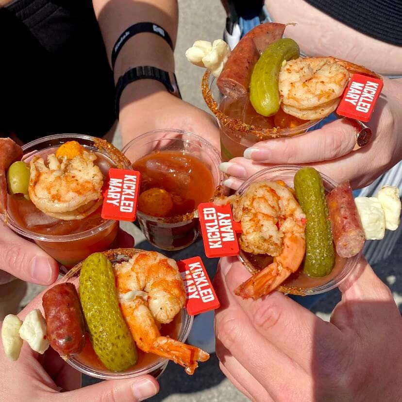 Five mini Bloody Mary cups held in a Cheers. The garnish includes a shrimp, a mini pickle, a part of a cocktail weenie, two cheese curds, and a Kickled Mary sign. One cup does not have a garnish.