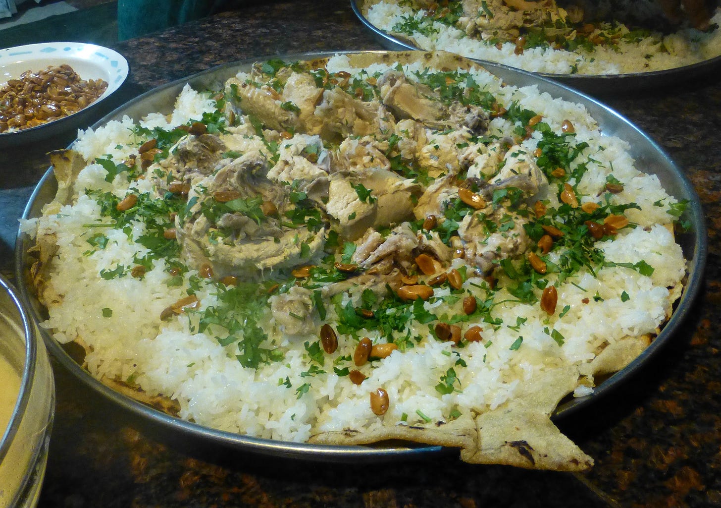 A large communal platter of mansaf: rice topped with pieces of meat, toasted pine nuts, and fresh parsley, served over flatbread. Additional side dishes are visible around the platter.