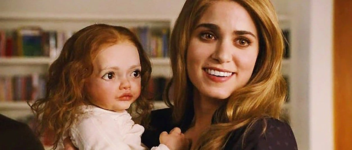 a atriz nikki reed, com cabelos loiros, pele branca, sobrancelha bem marcada e um olho cor de mel, segurando um bebê pouco realista de cabelo comprido e olhos esbugalhados.