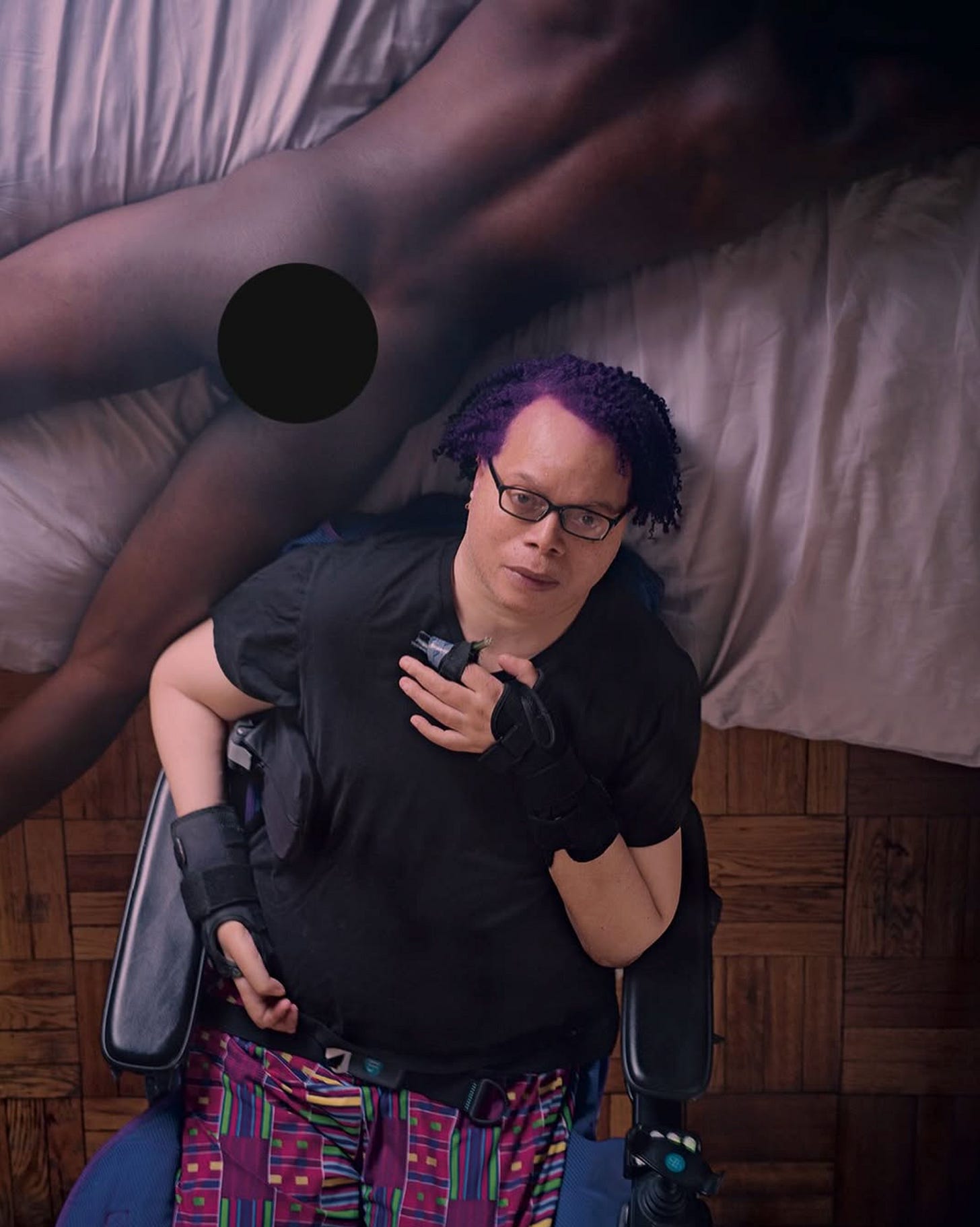 A photo taken from above of a Black disabled man with purple hair in a powerchair near a bed where a naked Black man is lying facedown with a black circle censoring his butt.