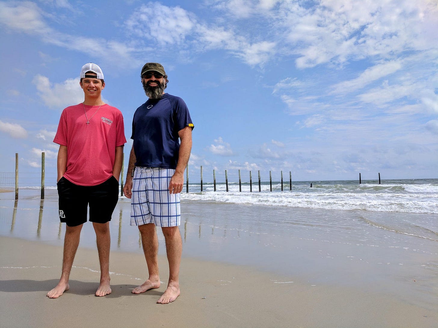 The author stands barefoot on a sandy beach beside his adult son. Both are smiling casually, dressed in shorts and T-shirts, with ocean waves rolling in behind them. Weathered wooden posts extend into the water along the shoreline under a wide blue sky with scattered clouds, evoking a calm, reflective moment shared between father and son. The author stands barefoot on a sandy beach beside his adult son. Both are smiling casually, dressed in shorts and T-shirts, with ocean waves rolling in behind them. Weathered wooden posts extend into the water along the shoreline under a wide blue sky with scattered clouds, evoking a calm, reflective moment shared between father and son.
