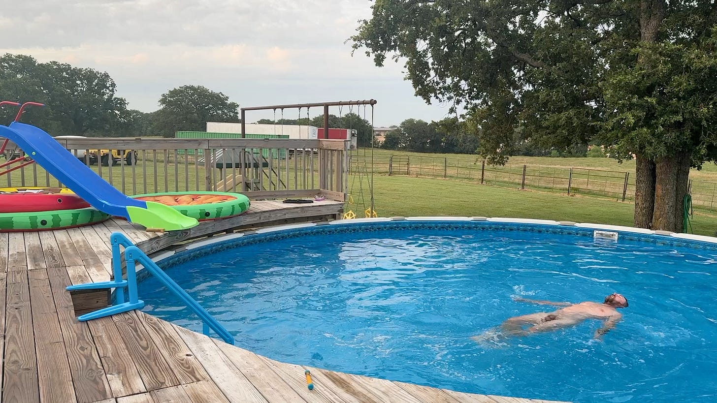 A nude man, Joel, floats on his back in a round above-ground pool surrounded by a wooden deck, slides, and open grassy farmland. A nude man, Joel, floats on his back in a round above-ground pool surrounded by a wooden deck, slides, and open grassy farmland.