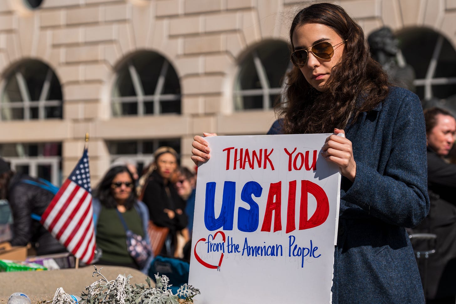 A woman among a crowd of supporters holds a sign thanking USAID employees in front of the Ronald Reagan Building in Washington, DC. A woman among a crowd of supporters holds a sign thanking USAID employees in front of the Ronald Reagan Building in Washington, DC.