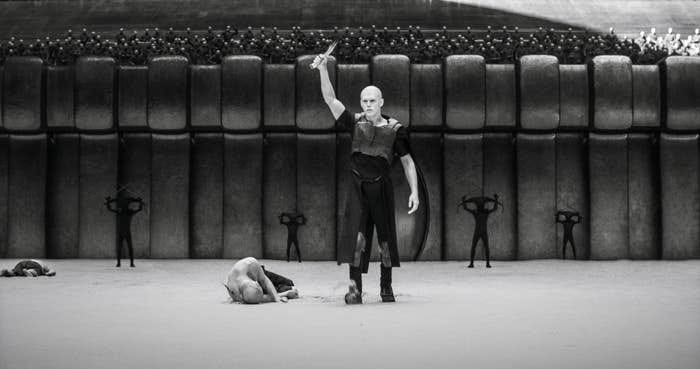 Person in a stylized pose on a stage with theatrical backdrop and figures in the background