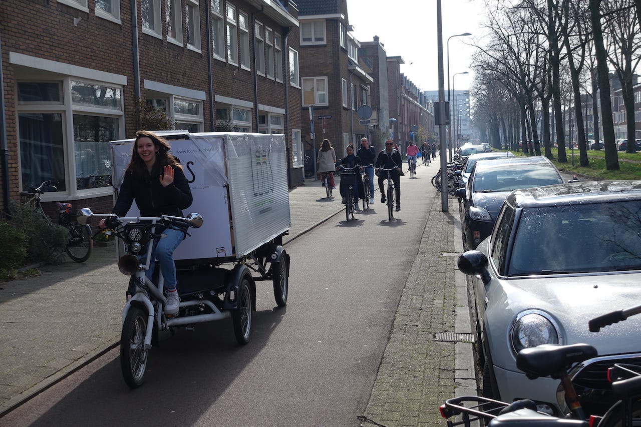 The tricycle and trailer are travelling along a cyclepath in Utrecht. The woman who was serving at the shop is now riding the tricycle and waving at the camera. The tricycle and trailer are travelling along a cyclepath in Utrecht. The woman who was serving at the shop is now riding the tricycle and waving at the camera.