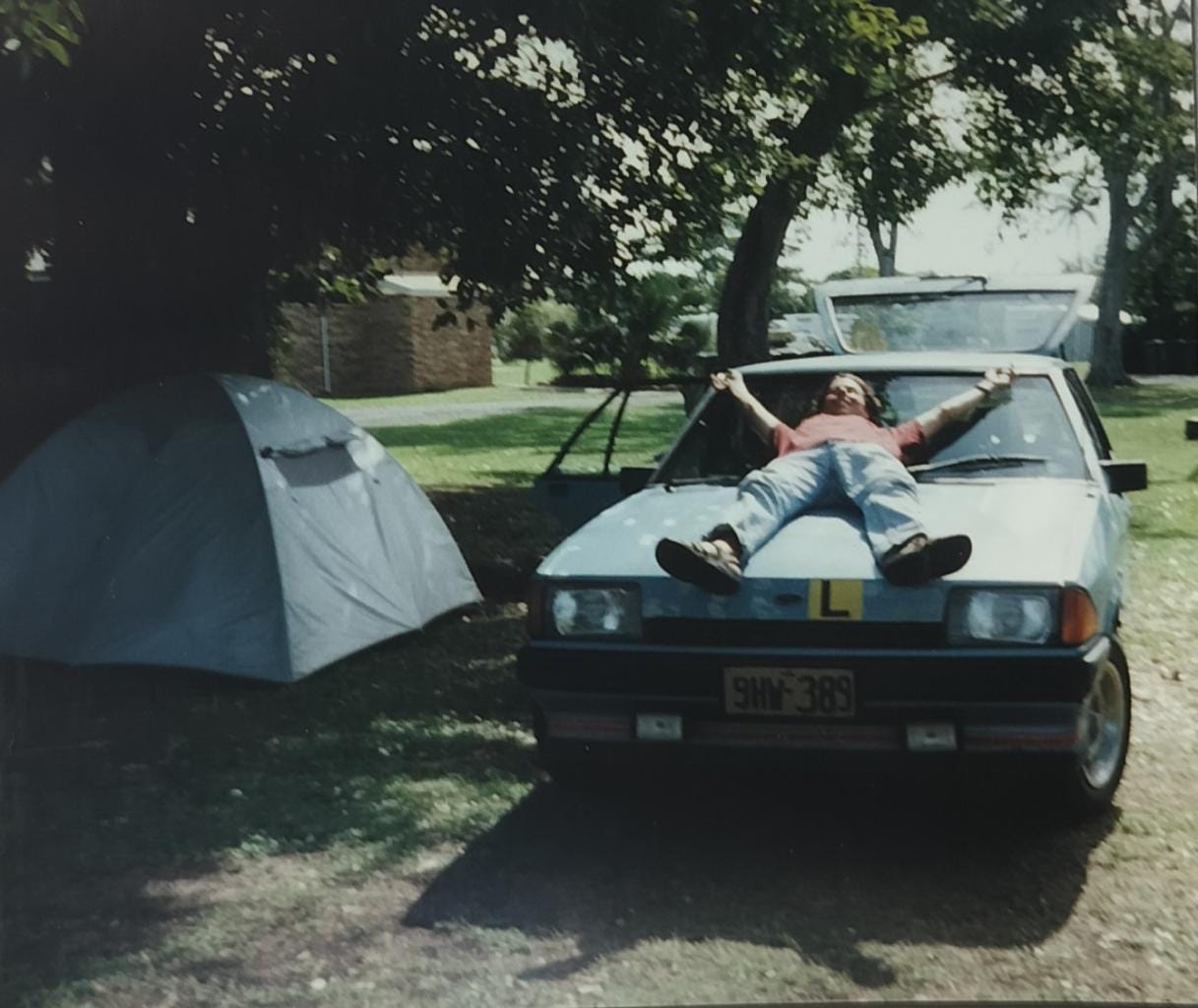 A young man lying on the bonnet of a beat-up Ford Falcon next to a tent at a campsite in Australia
