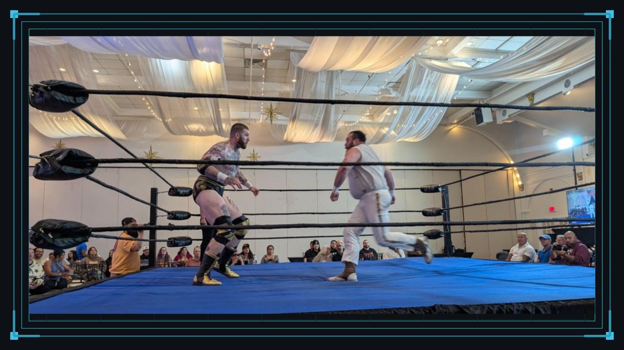 Two professional wrestlers move toward one another in the center of a blue wrestling ring. The ring is surrounded by spectators, with black ropes crossing the foreground and white drapery and string lights covering the venue ceiling.