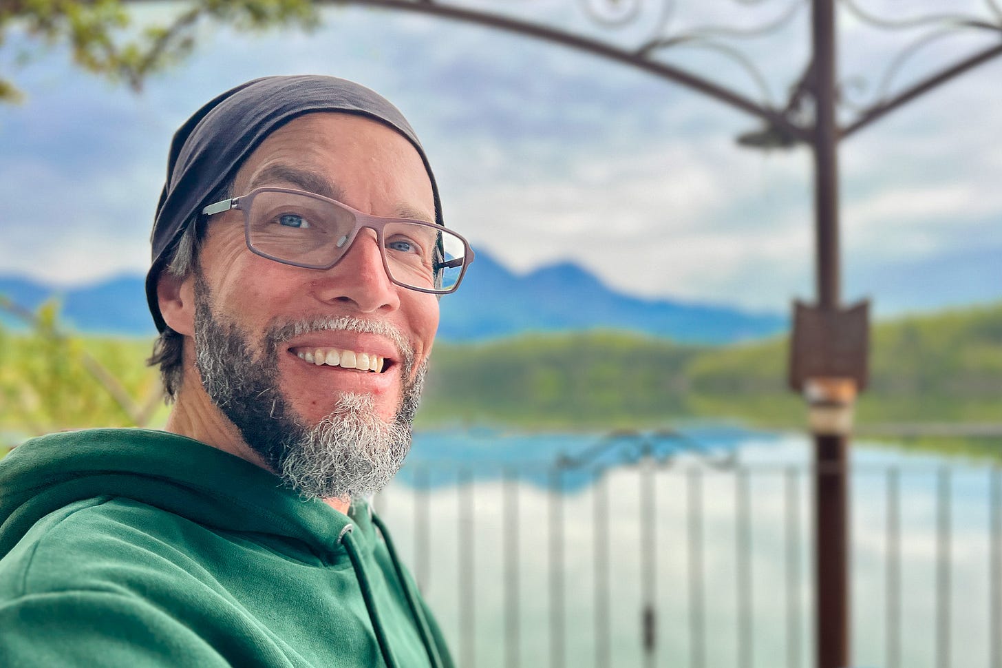 Patrick Hemminger mit Brille, Bart und dunkler Mütze sitzt im Freien vor See und Bergkulisse. Er trägt einen grünen Hoodie und blickt zur Kamera; im Hintergrund sind Geländer, Wasser, bewaldete Hänge und ein wolkiger Himmel zu sehen.