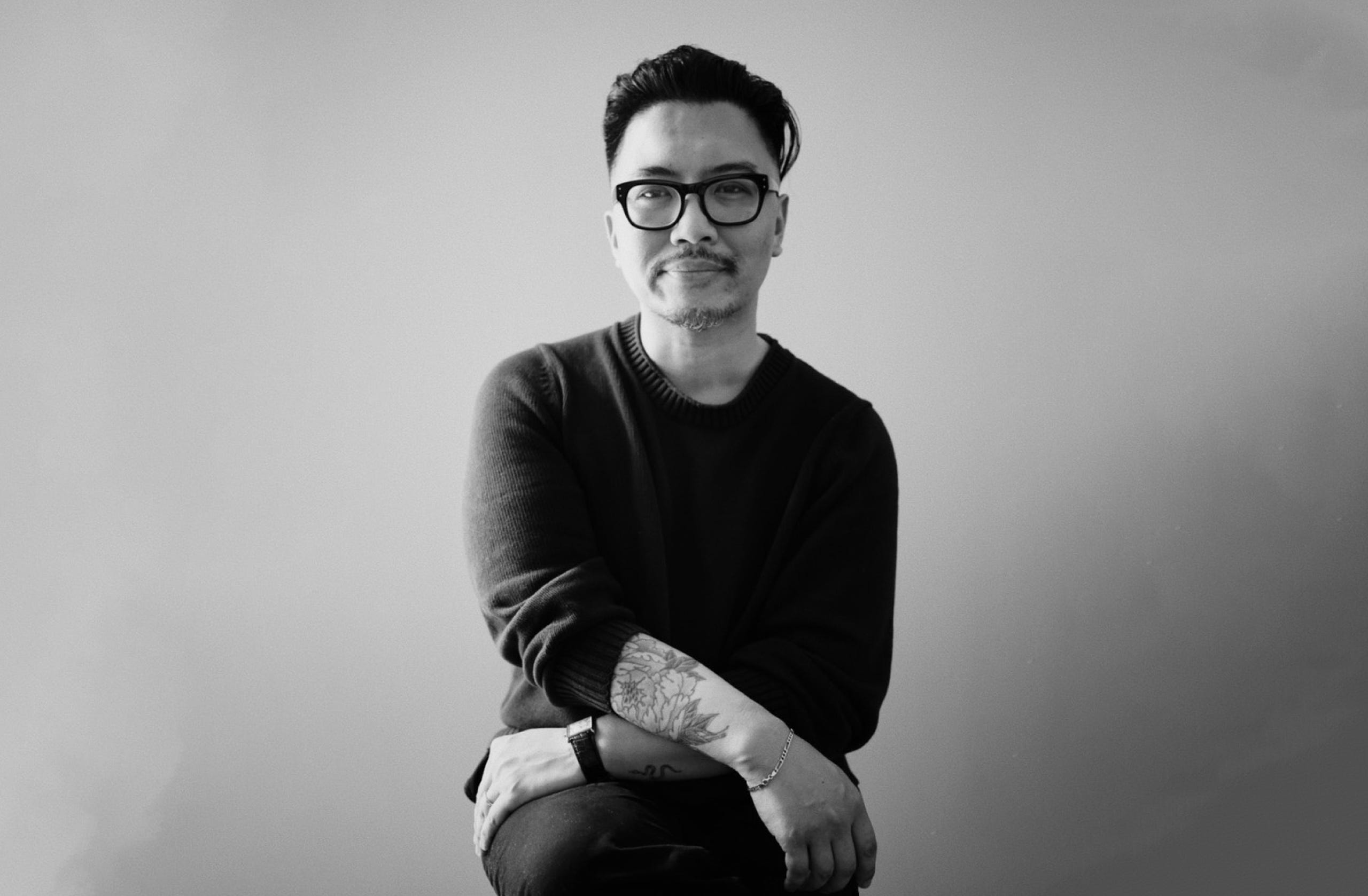 Peter Nguyen sits facing the camera with his arms crossed on his lap posing in black and white.