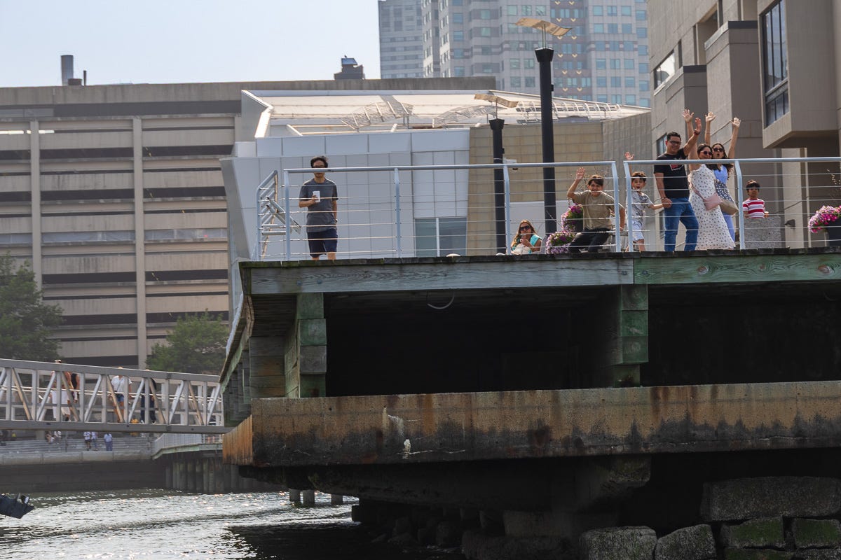 Group of people waving from a wharf in Boston Harbor as a boat passes