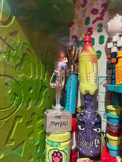 First picture shows a jumble of colorful items agains a green wall. One of the items is a trophy labeled Sports Ball. 2nd picture a young man and a man with a white beard sitting in front of two round windows. There is a green steel girder separating the sitters. #rd pic: The same two people are sitting on a couch in front of shelves with books piled haphazardly.