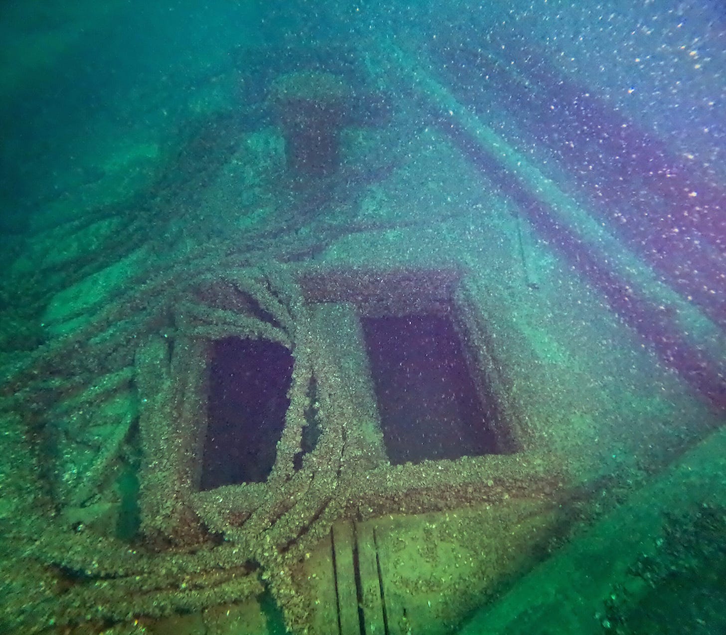 Underwater view of a shipwreck with two square openings and ropes covered in marine growth, surrounded by greenish-blue water.