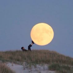 This may contain: two people are standing on top of a hill with the moon in the background