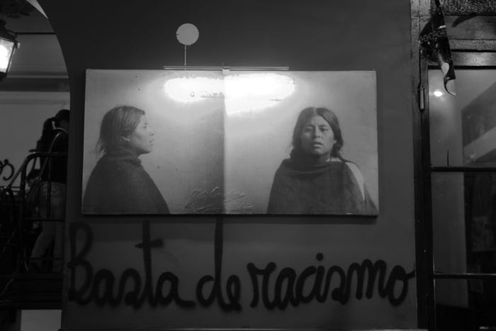 Black and white photo of dimly lit street. On the wall facing us there are two photos of the same woman, front and side angle: it looks like a police mug shot. Scrawled in black cursive graffiti is the slogan "Basta de racismo". It is unclear what circumstances would have led to this woman’s apprehension. She is likely from an Indigenous tribe. This image of the photos and grafitti is used by the anarcho-feminist group Mujeres Creando Feminismo in Bolivia, who use grafitti as a method of political expression.