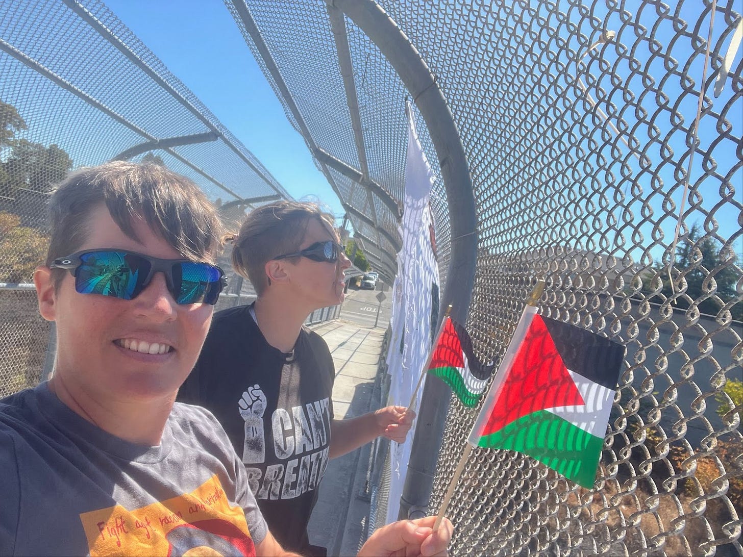 selfie of me and my partner wearing sunglasses and waving small palestinian flags at cars driving by on a highway overpass.