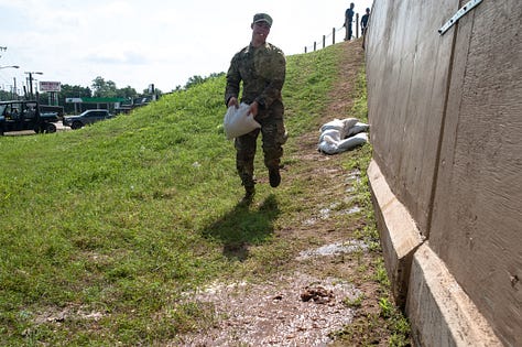 The progression of a sand boil from occurrence to remediation: 1) A soldier in fatigues carries a sandbag downhill alongside a floodway structure (a tall wall) with a cigarette between their lips and eyes turned down toward the target in the grass. The sand boil looks like a gurgling fountain a few inches in diameter, with muddy water spreading around it. 2) The floodway structure, chain of soldiers and ring dyke (several dozen sandbags stacked around the sand boil in a half-moon formation at the base of the wall) are seen from just uphill. A person in a neon yellow safety vest stands in muddy, ankle-deep water as it rises inside the ring dyke, which is now a few sandbags high. Inside the floodway structure, floodwaters as sludgy as brownie mix appear to loom several feet over the heads of the soldiers working on the landside. Two military SUVs pass on the road in the background. 3) Soldiers in fatigues pass sandbags down from pallets on a flatbed. In the foreground, a blond soldier with a square jaw and solemn expression looks on in a camouflage cap. 4) Soldiers in fatigues stand perpendicular to the floodway structure, passing sandbags toward the ring dyke. 5) Two people stand side by side, facing the ring dyke with their backs to the photographer. Muddy water spills over the sides of the ring dyke, which now stands five or six sandbags high. The person on the right is in fatigues. The person on the left wears a neon yellow safety vest with the red and white turreted castle emblem of the U.S. Army Corps of Engineers. 6) Several members of the Oklahoma National Guard stand in, on and around the ring dyke in fatigues. One is submerged to calf- or knee-height in the muddy water spilling over the edges of the ring dyke. Another steps calf-deep into the pool with one leg, straddling the ring dyke to position a sandbag.