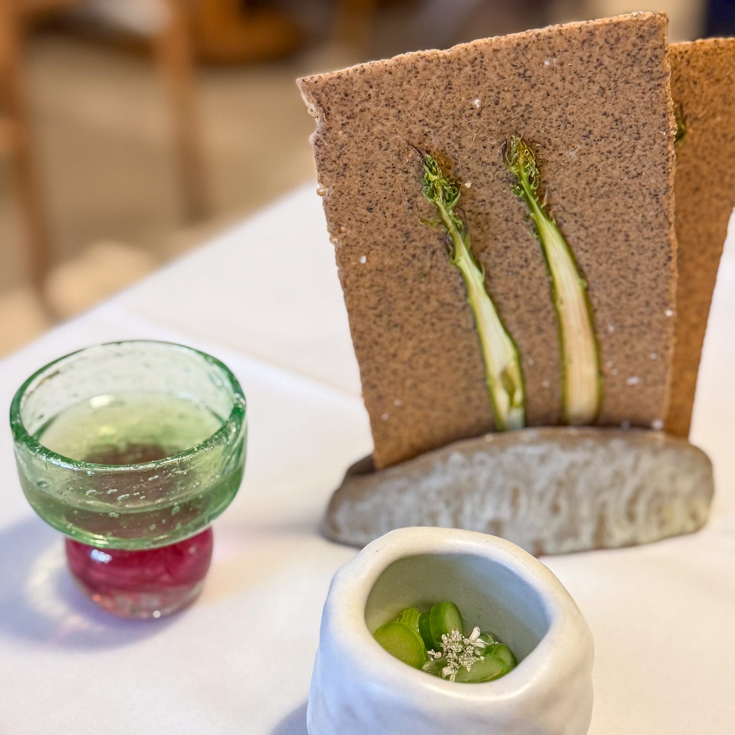 Asparagus-skin consommé in a green glass, a speckled asparagus tuile, and a small pot of sliced asparagus with white blossom, at Datil in Paris. Asparagus-skin consommé in a green glass, a speckled asparagus tuile, and a small pot of sliced asparagus with white blossom, at Datil in Paris.