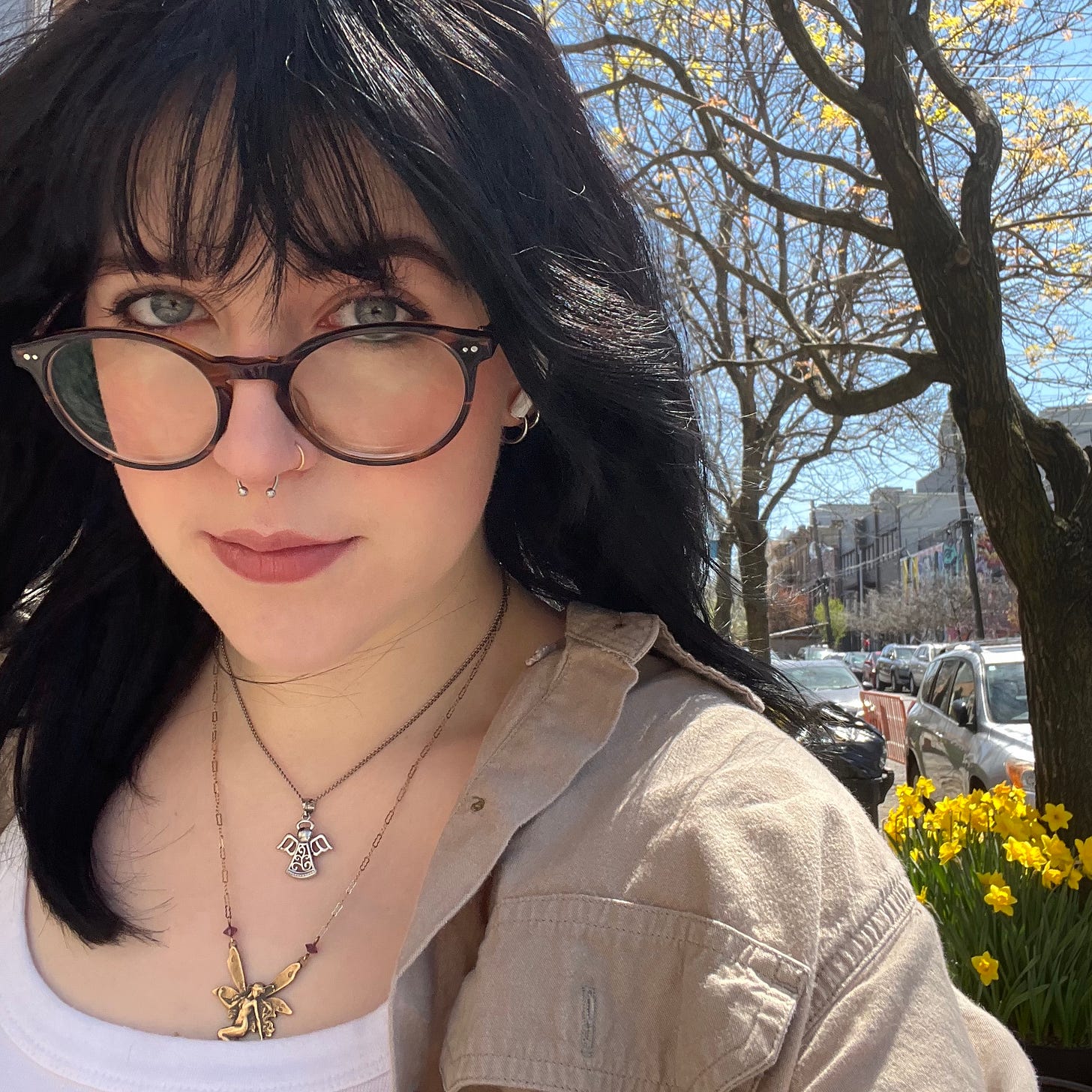 selfie of June on a sunny day, wearing a white tank top and beige button down shirt and two necklaces. daffodils are pictured behind her left shoulder in a tree pit on the sidewalk