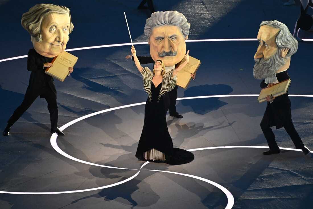 Italian actress Matilda De Angelis (center) performs as a conductor, dressed in a black gown and holding a large baton, with dancers dressed as the three great masters of Italian opera Giuseppe Verdi, Giacomo Puccini and Gioachino Rossini, who are wearing giant heads and holding books of sheet music. Italian actress Matilda De Angelis (center) performs as a conductor, dressed in a black gown and holding a large baton, with dancers dressed as the three great masters of Italian opera Giuseppe Verdi, Giacomo Puccini and Gioachino Rossini, who are wearing giant heads and holding books of sheet music.