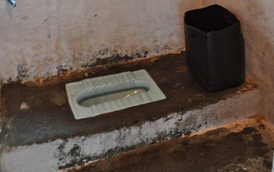 White porcelain squat toilet, surrounded by discolored cement and a black bucket beside it. White porcelain squat toilet, surrounded by discolored cement and a black bucket beside it.