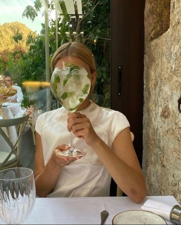 This may contain: a woman sitting at a table holding a wine glass in front of her face with water inside