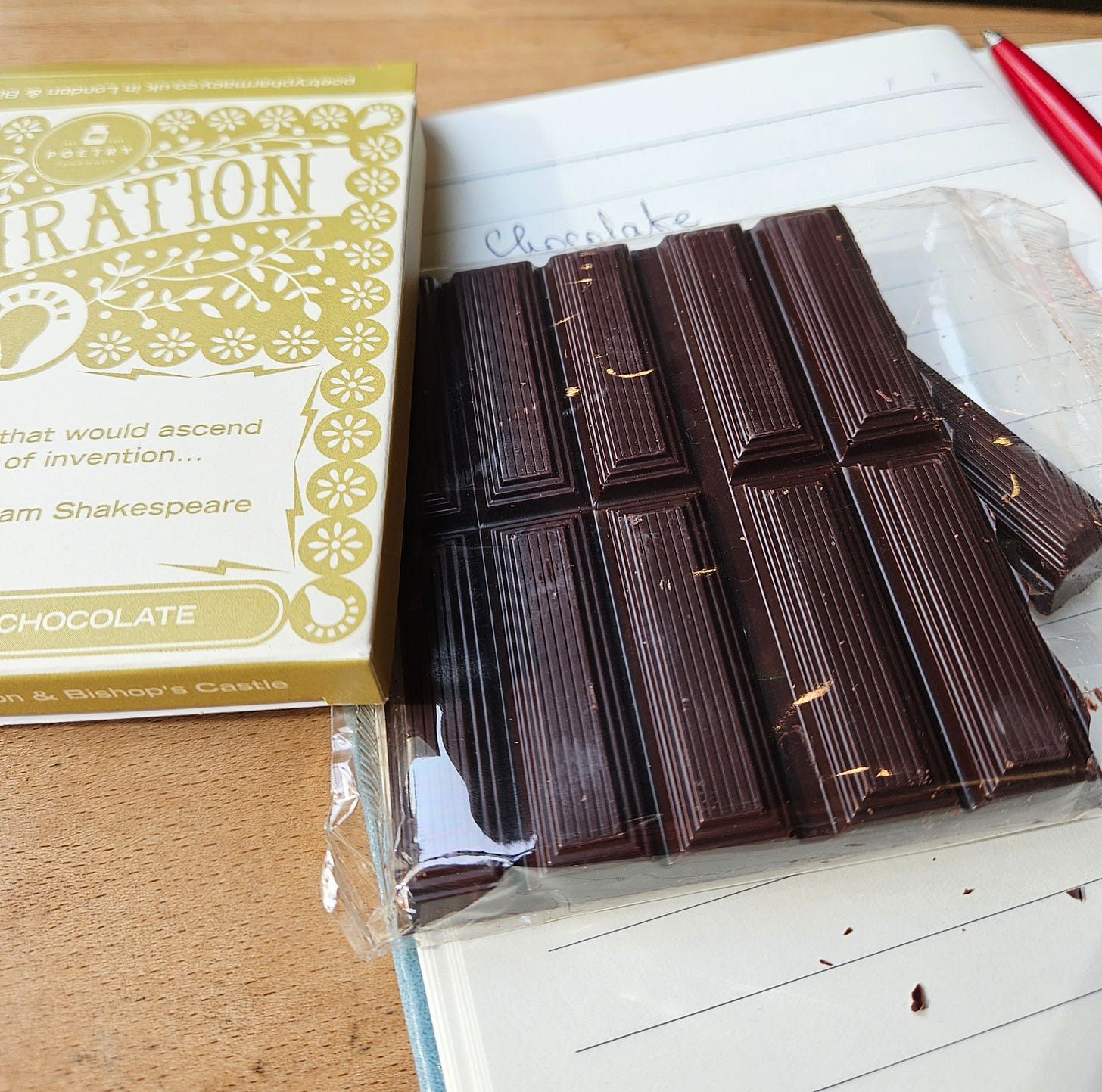 The image shows a partially opened package of dark chocolate. The chocolate is dark brown and divided into rectangular segments with a textured surface. Next to the chocolate is a yellow and white box with decorative floral patterns. The text on the box reads "INSPIRATION" at the top. The chocolate is resting on a notebook or piece of paper with handwritten text that appears to say "Chocolate."