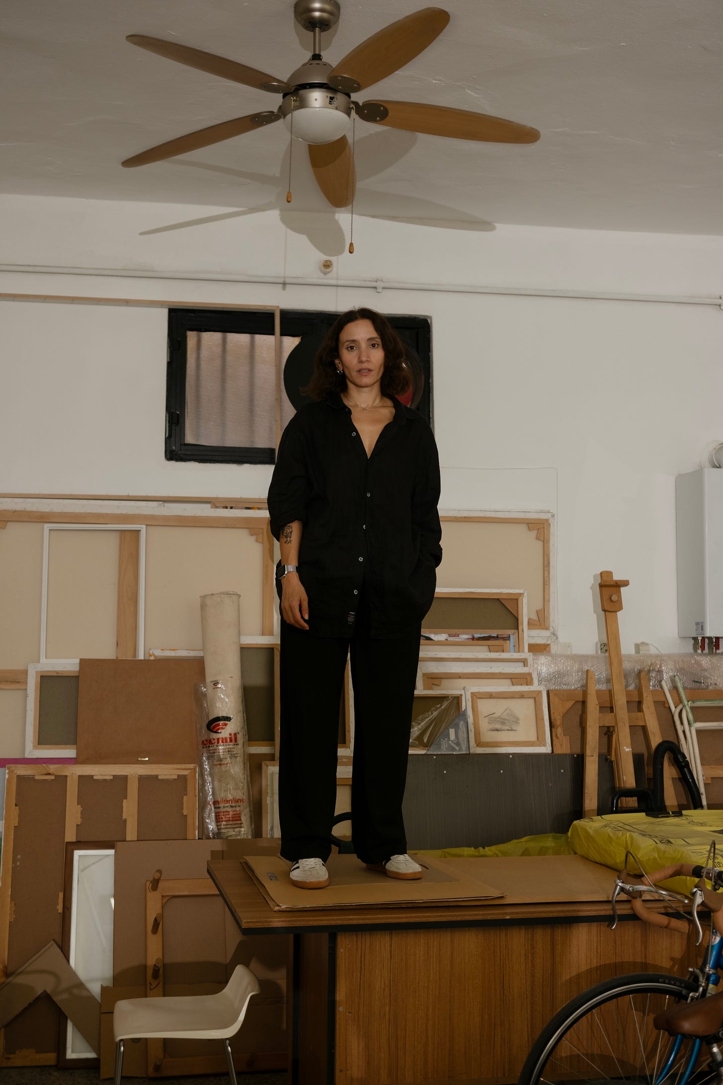 Full-length photo portrait of the Turkish artist Merve Mepa, a woman with shoulder-length dark hair in a black shirt and black trousers, standing on a desk under a rotary ceiling fan, in the middle of a crowded studio with unfinished canvas and frames leaning against the wall behind her.