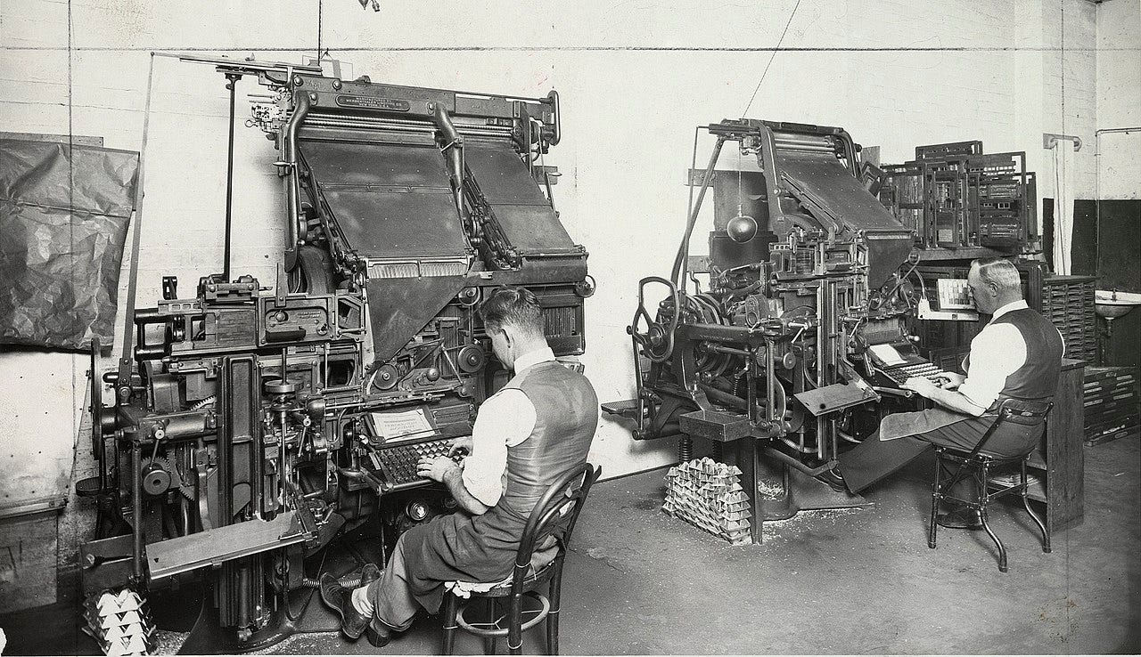 Black-and-white photo of two men seated at linotype machines in a large office. The machines and very tall, made of metal, and have a lot of moving mechanisms. Black-and-white photo of two men seated at linotype machines in a large office. The machines and very tall, made of metal, and have a lot of moving mechanisms.