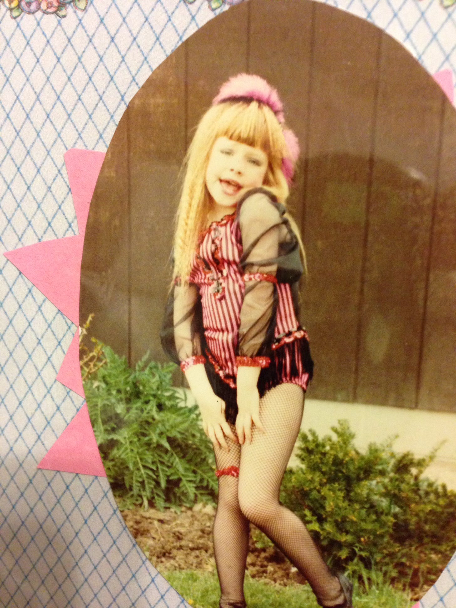 A white child wears a fringed black and pink costume with fishnets and a garter around her leg, and she is making an uncomfortable "sexy" pose.