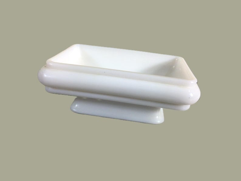 May include: A white ceramic soap dish with a raised rim and a base. The dish is rectangular and has a smooth, glossy finish.