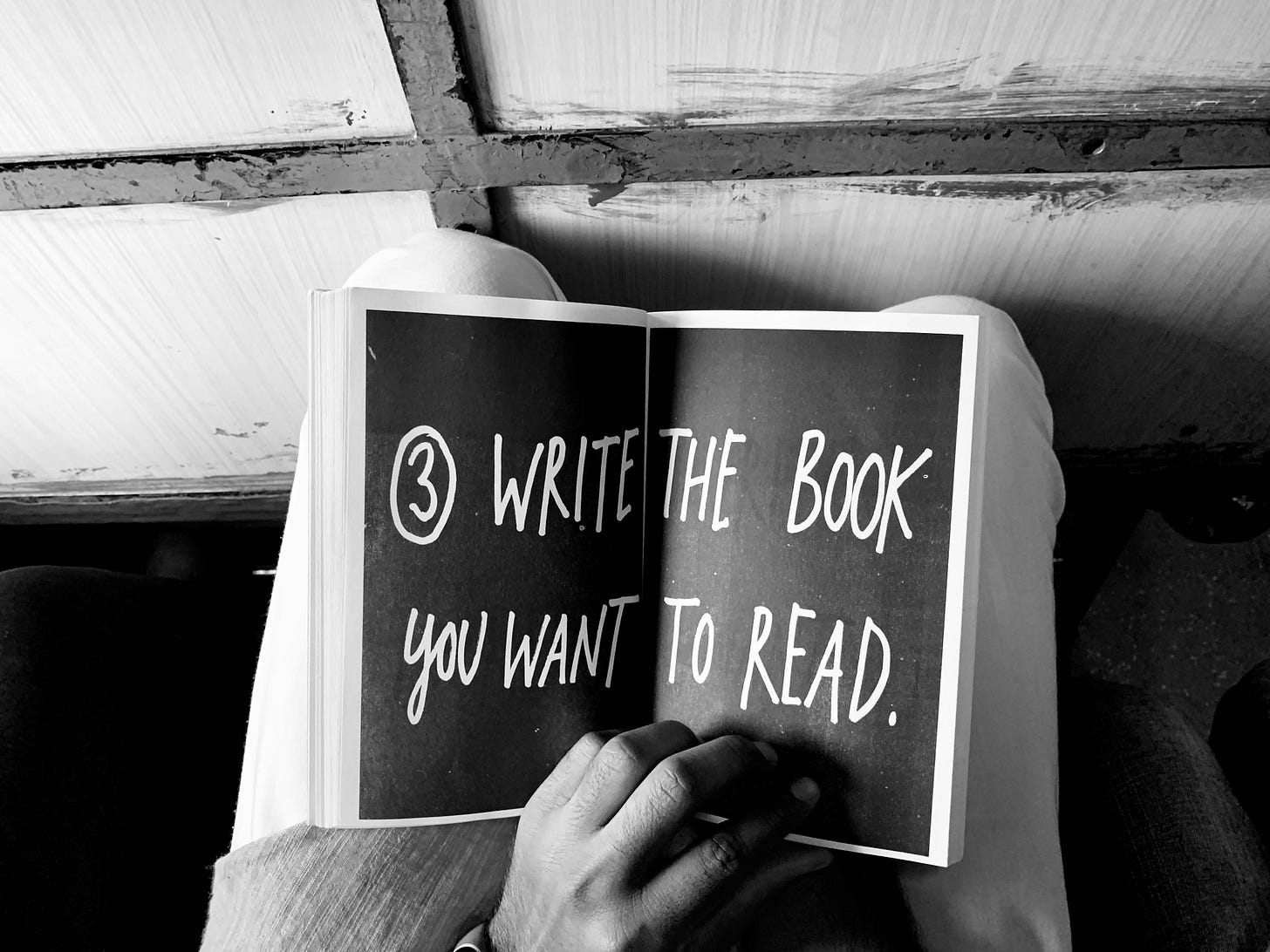 An open book on someone's lap that says "Write the book you want to read" in white font on a black background across a 2-page spread. An open book on someone's lap that says "Write the book you want to read" in white font on a black background across a 2-page spread.