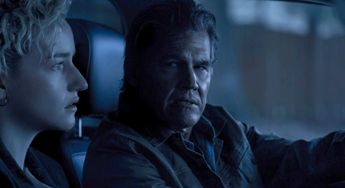 Mulher e homem sentados dentro de um carro, iluminados por uma luz fria e azul; ambos com expressões sérias e tensas. Mulher e homem sentados dentro de um carro, iluminados por uma luz fria e azul; ambos com expressões sérias e tensas.